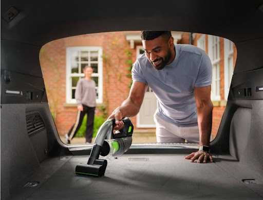 Man vacuuming a car boot with a Gtech Multi handheld vacuum to remove dirt, grit, and pet hair from the carpet.